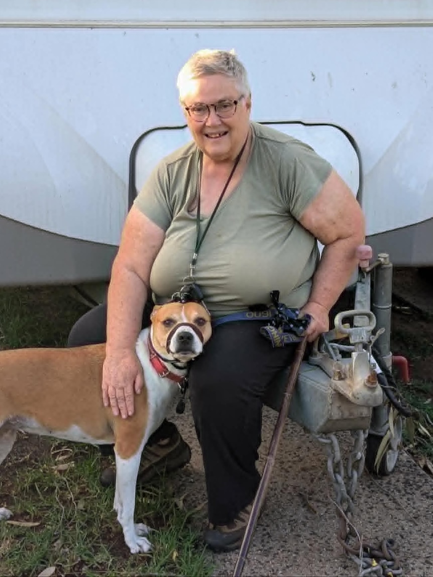 A woman sits in front of her caravan while holding her brown and white dog.