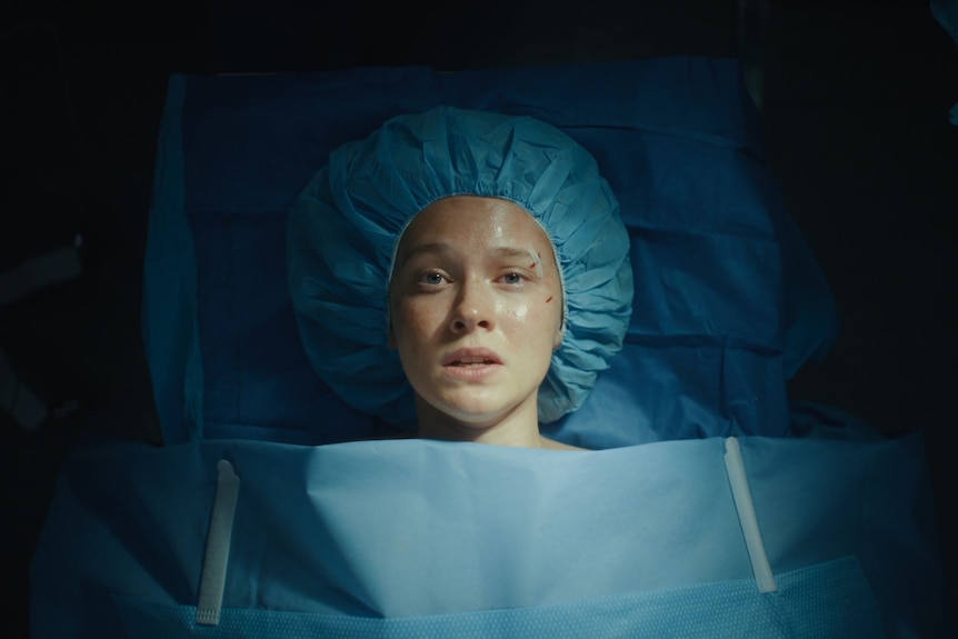 A young woman with a butterfly stitch on her eyebrow and wearing a blue scrub cap, looking panicked on a hospital bed.