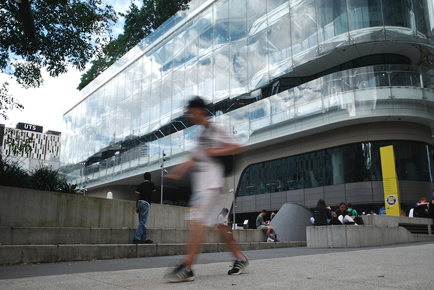 A person carrying a bag walks through a university campus. Behind them is a large multi-story glass building. 