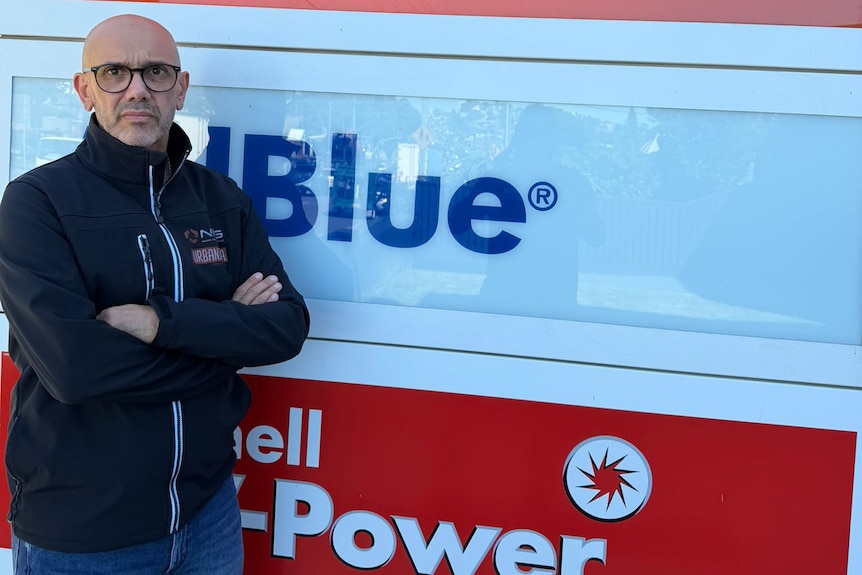 A bald man with dark rimmed glasses in a navy jumper stands, staring ahead with his arms folded beside a service station sign.