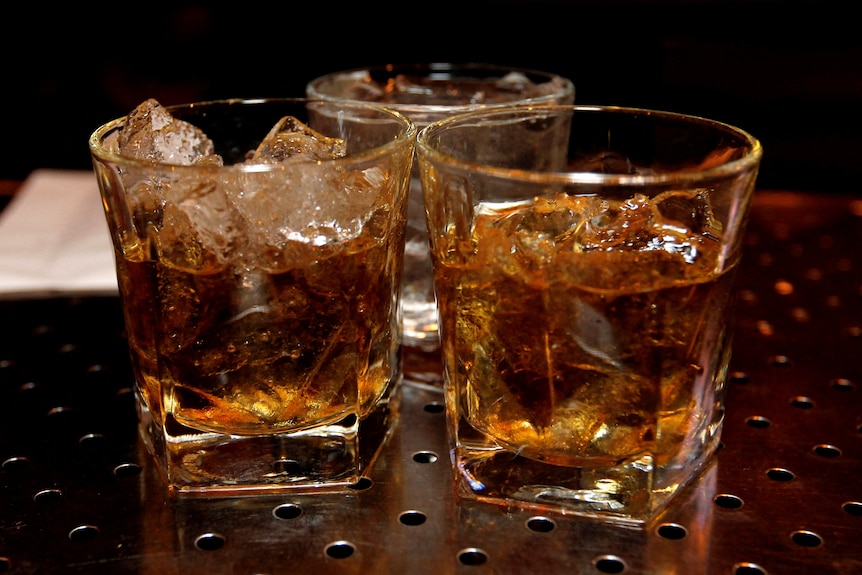 Glasses of whiskey on ice sit on a bar.