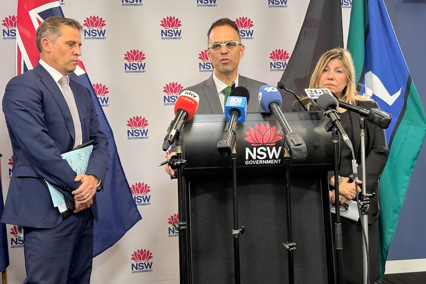 Mr Mookhey standing at a lecturn speaking to media into microphones in front of a NSW government backdrop.
