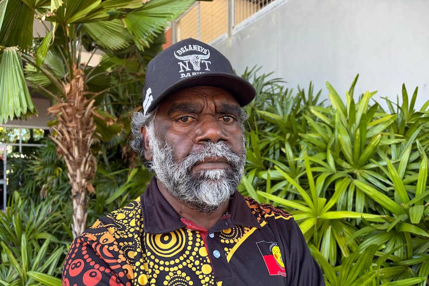 Man wearing polo with Indigenous art print looks at camera standing in front of green plants