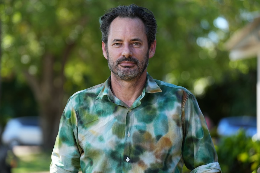 A man wearing a patterned button-up shirt stands in a park, greenery and trees can be seen in the background.