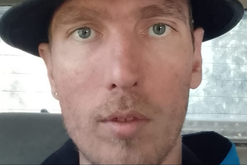 A man in a dark hat sits in a car and takes a selfie.