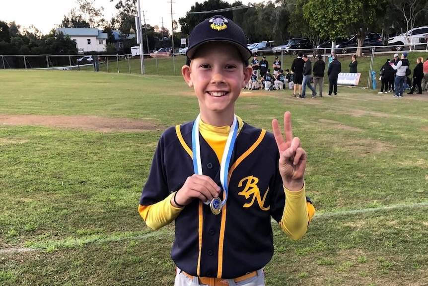 A young boy gives a  peace sign while showing off a medal on a baseball field