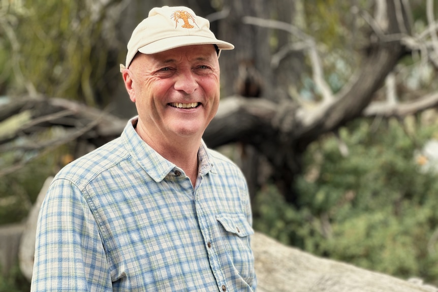 An older man wearing a cap with forest in the background.