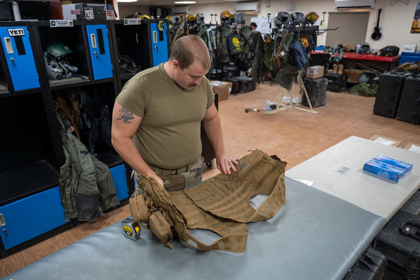 A soldier inspects a survival vest laid out on a table. He is in khakis. Other equipment behind.