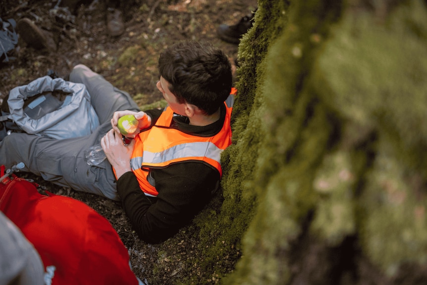A male wearing an orange hi-vis vest over his clothing leans against a mossy tree. He holds an apple
