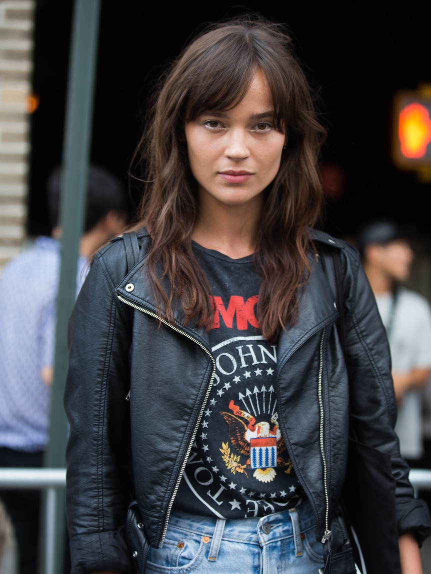 A woman standing outdoors on a city street wearing a leather jacket and Ramones t shirt, staring at the camera.