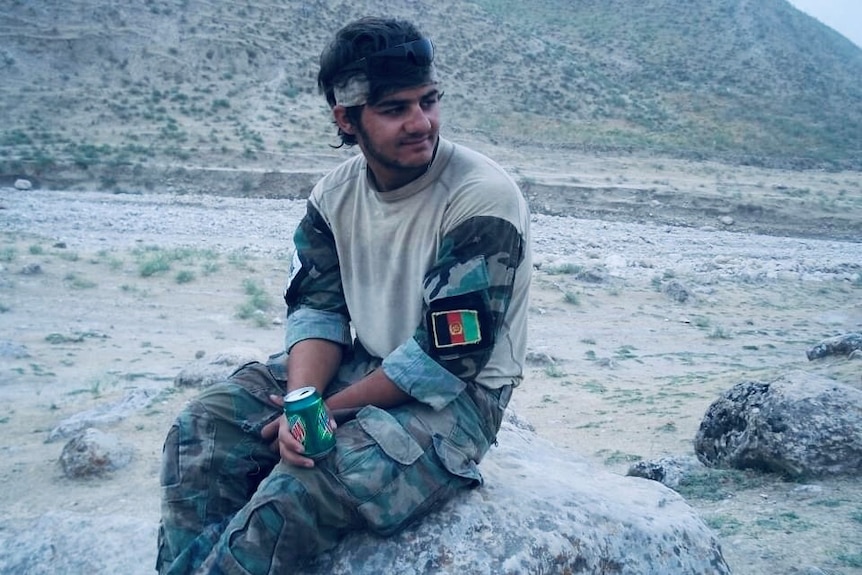 A man wearing camoflague pants and shirt sits on a rock, smiling.