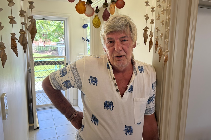 An older, grey-haired man stands in the hallway of his house.