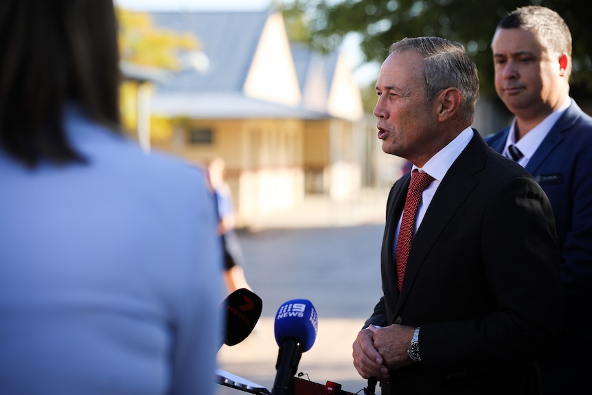 A side-on shot of WA Premier Roger Cook speaking outdoors at a media conference with a man in the background.