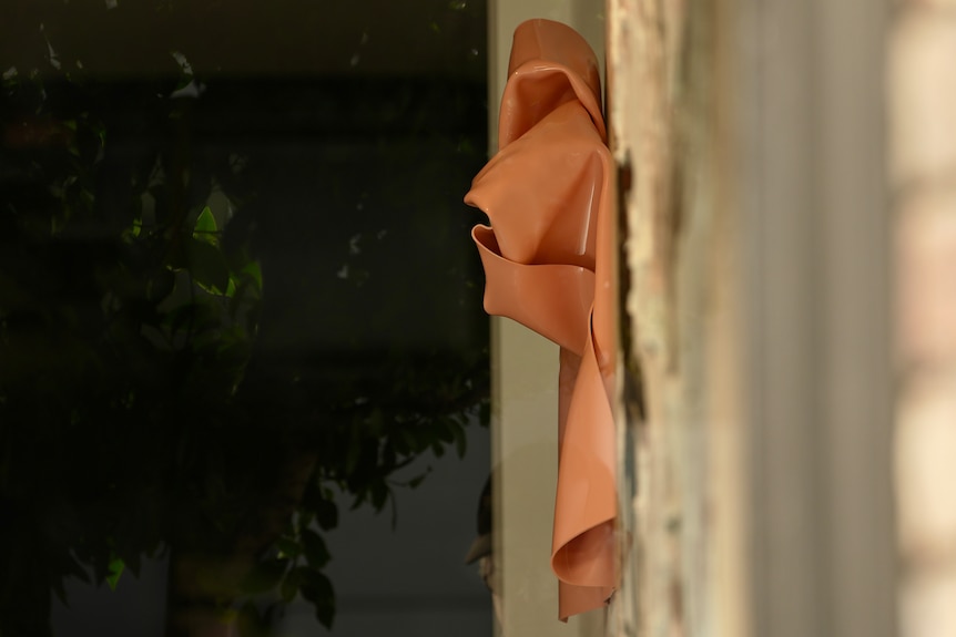 An orange plastic sculpture that looks like a crumpled piece of cloth hangs on the wall of a gallery.