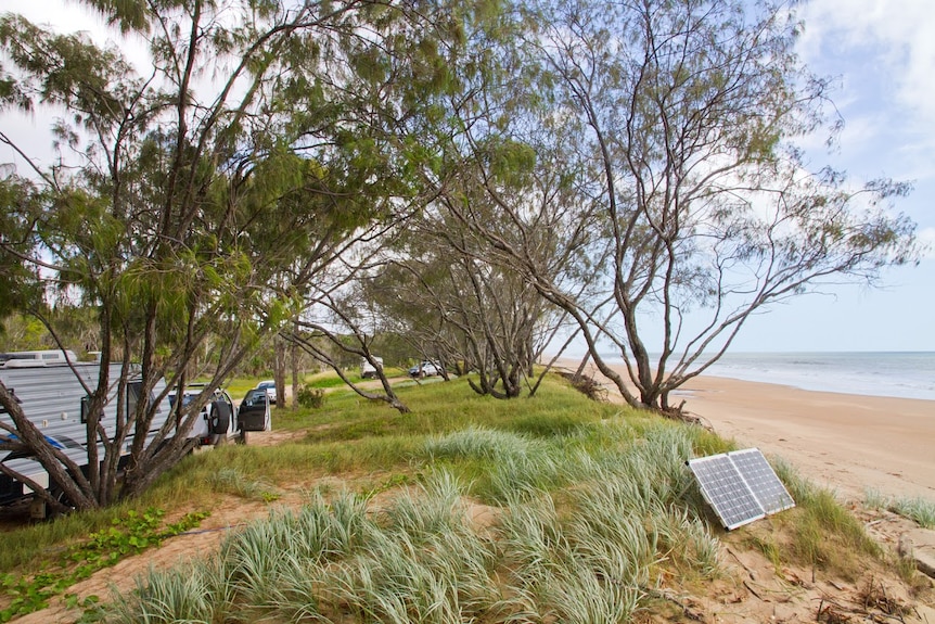 Caravans, vehicles and a solar panel are dotted along a sand dune beside a beach.