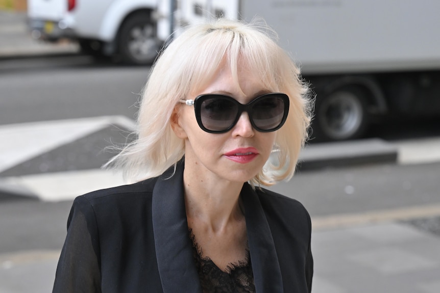 A woman with blonde hair and large black sunglasses in front of a road. 