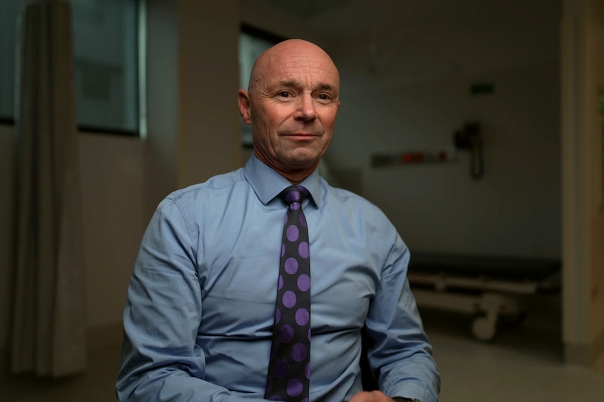A bald man wearing a blue business shirt and purple patterned tie sits in an empty hospital ward.