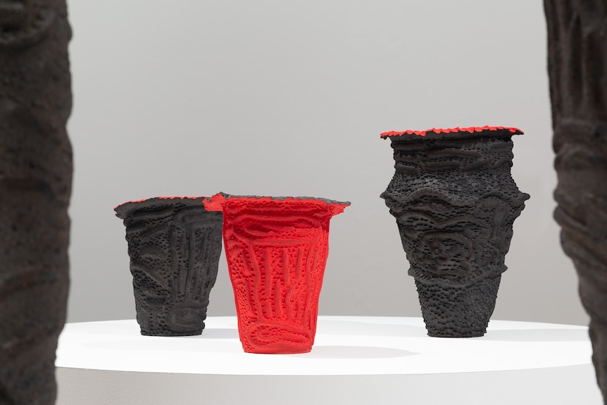 Three textured urns stand on a platform in a white gallery. Two are painted black, the one in the middle is red.