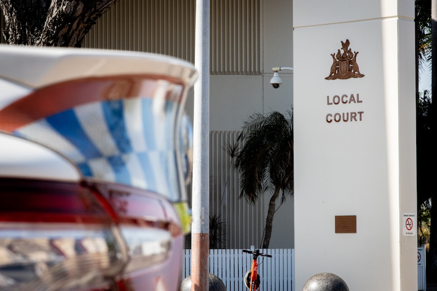 The boot of a police car blurred in the foreground, with the local court entrance sign in the background in focus.