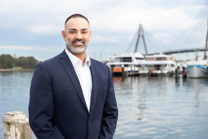 A man wearing a white shirt and dark blue suits stands in from of a fishing fleet in a marina.