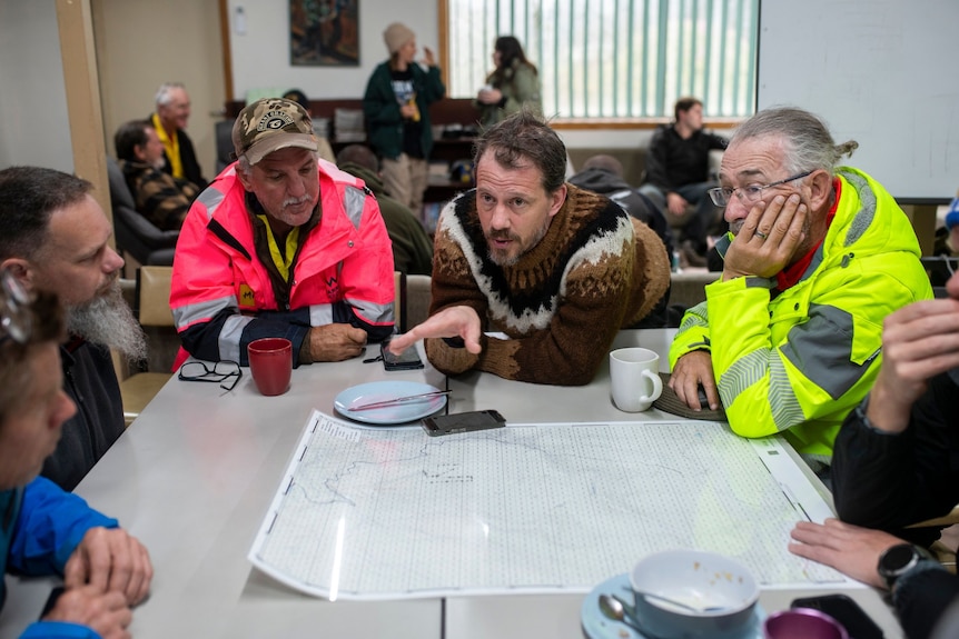 A group sits around a table with a large land map in front. Centre is a man speaking using hand to gesture.  Five others listen