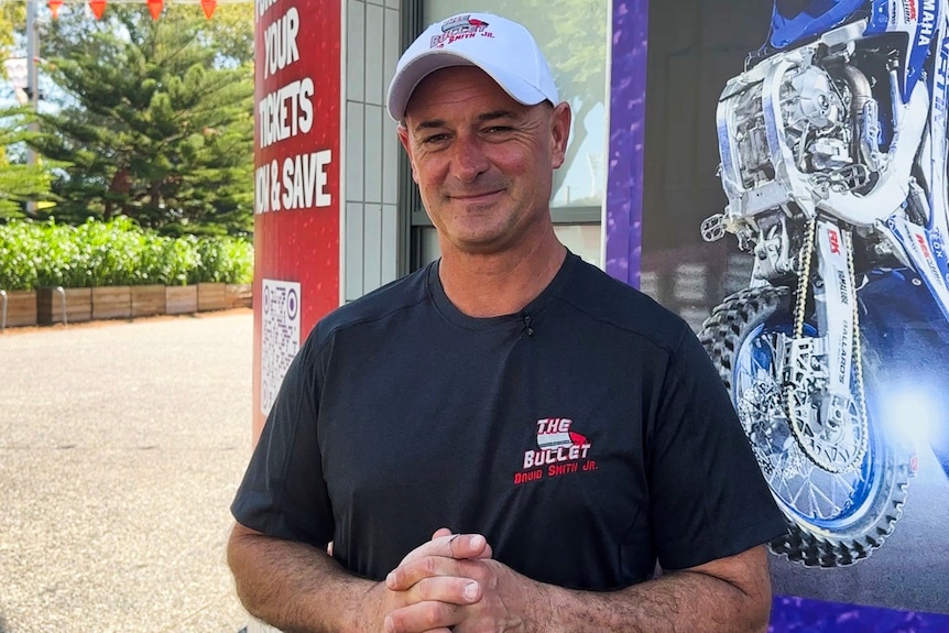 A man in a black shirt and white hat standing outside in front of a picture of a motorbike.