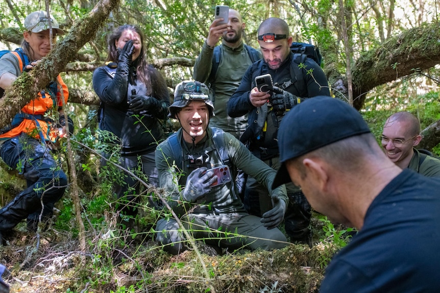 Six people in dark clothing bushwalking gear react in various happy ways looking at another man. They are in the forest