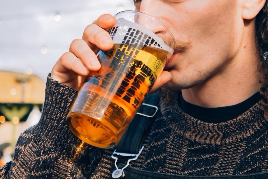 A close-up of a young person with facial hair and a mullet drinking an amber beverage that looks like beer.
