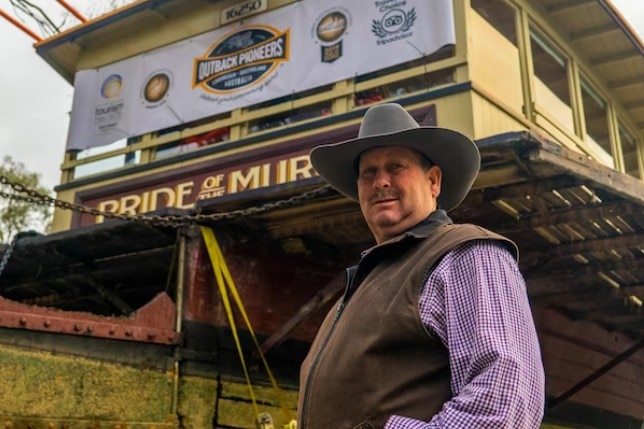 A man with a cowboy hat stands near an old boat