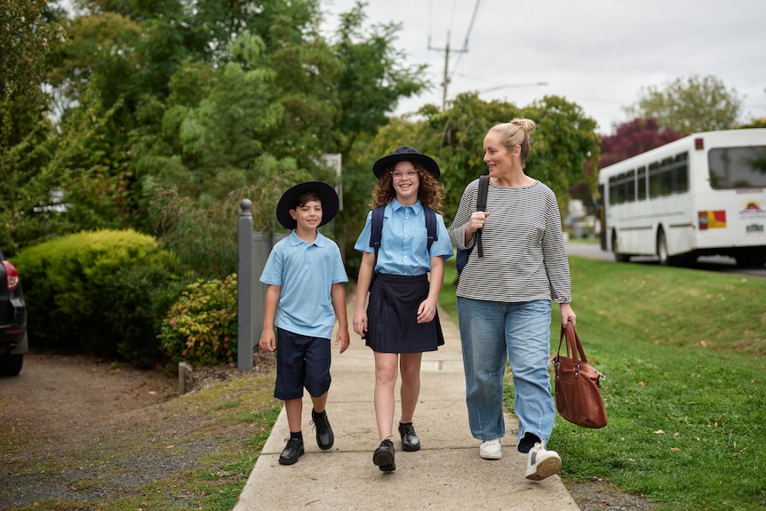 A mother wearing jeans and a grey shirt walks down the street with two children both in blue uniforms, one a girl, one a boy.