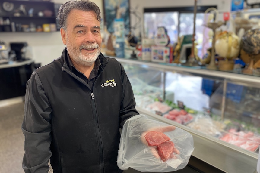 A man holding a tuna steak stands at a seafood store's counter, looking at the camera.