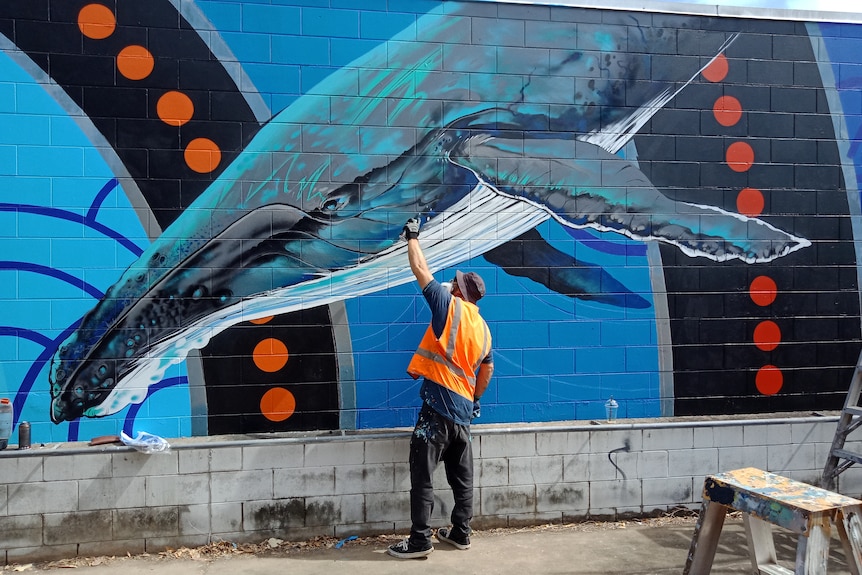A man in an orange hi-vis vest paints a whale on a wall.