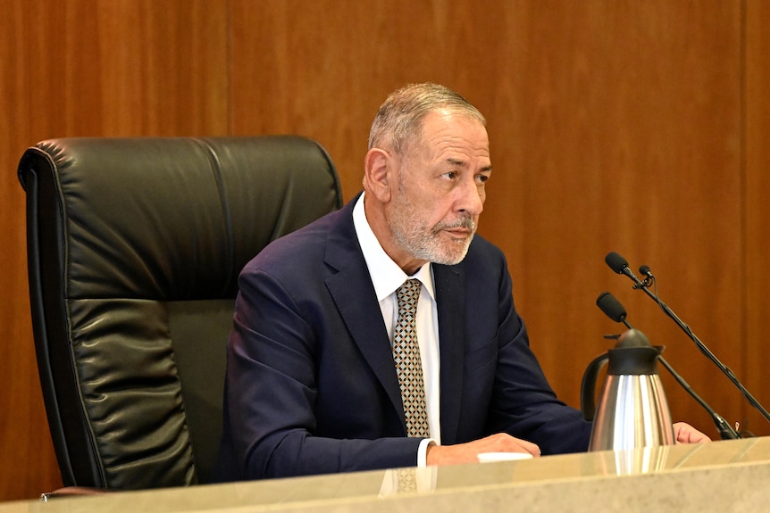 A man in a suit sitting in a high-back leather chair at a timber bar with microphones on it. 
