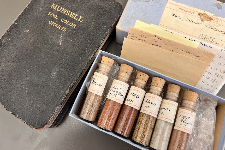 An old black book lays next to coloured soil samples on a silver bench.