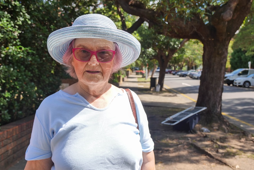 an older woman wearing a hat and pink glasses smiles gently