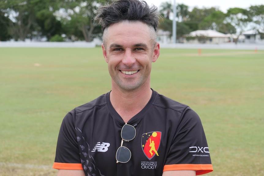 A man with messy hair smiling at the camera, a cricket pitch