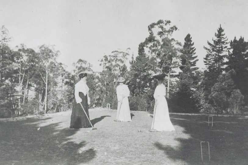 black and white photogrpah of women in white dresses on grass area, with hoops, balls an mallets surrounded by trees