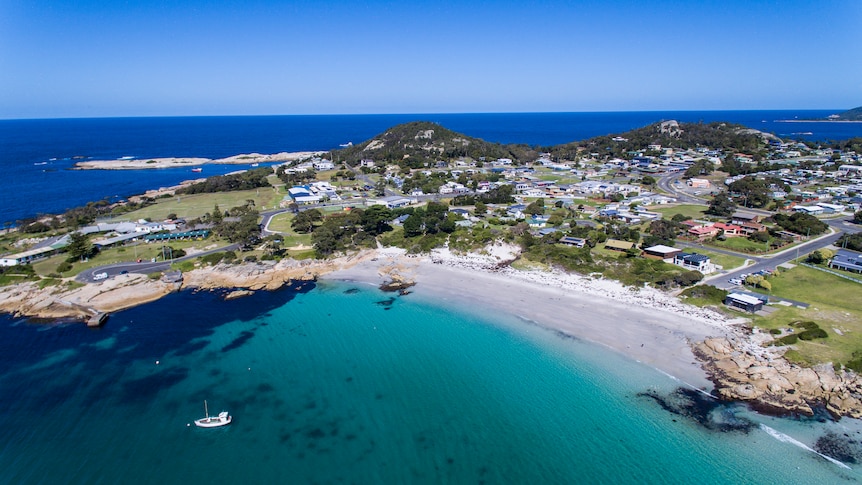 An aerial view of Bicheno showing houses and buildings on a headland with a beach in the foreground and water behind.