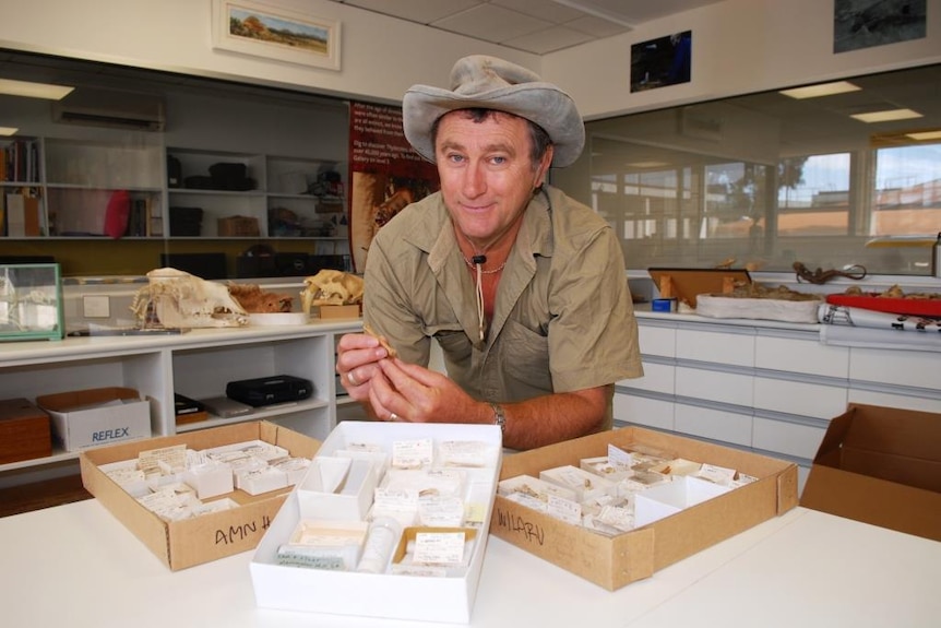 A man wearing a khaki shirt and hat smiling at the camera and holding a small bone