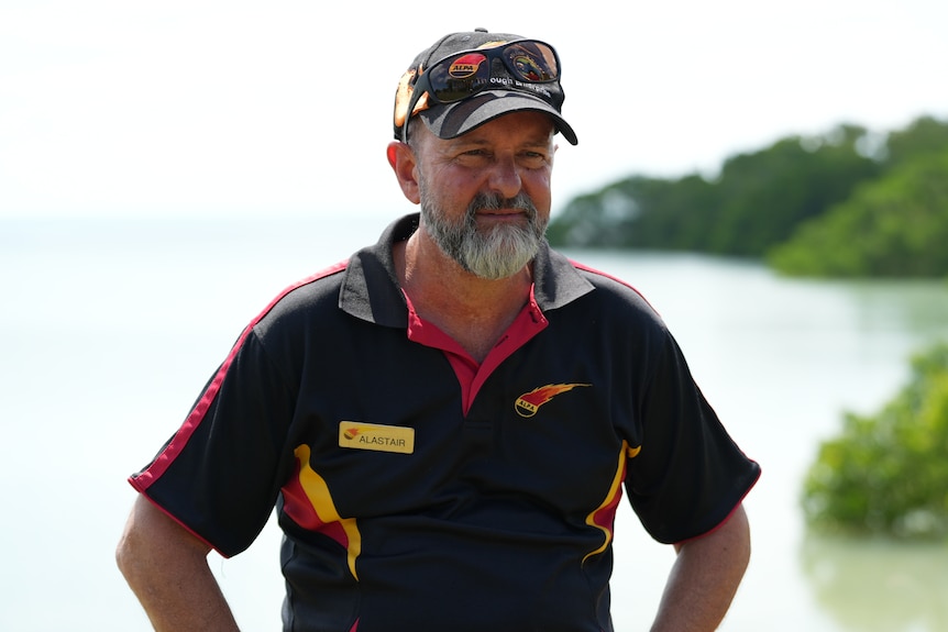 A man on the beach has a serious facial expression and is wearing a shirt displaying the Arnhem Land Progress Association logo.