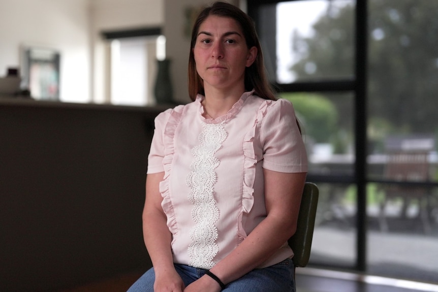 A woman in a pale pink top and pants sits straight in a chair in a kitchen and dining room, serious facial expression
