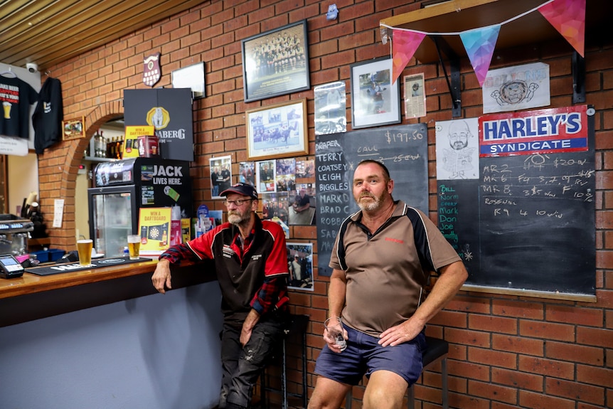 Man wearing grey work shirt sits stony-faced inside pub in front of chalkboard wall