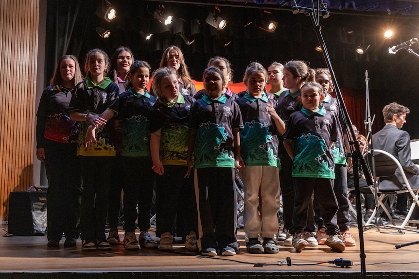 A dozen young children stand on stage in matching shirts