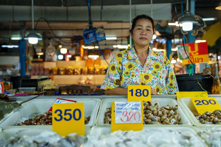 A woman wearing a shirt decorated with bright sunflowers looks glum, as she stands behind buckets of snails for sale.