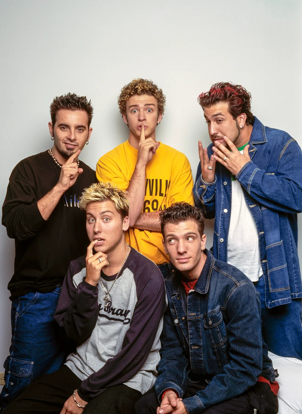 Five men posing playfully with fingers to lips, wearing casual 90s-style outfits including denim and graphic tees