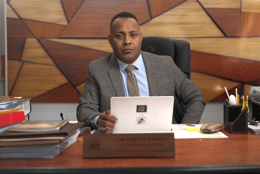 A man in a grey suit holds a white tablet while sitting in a wood-panelled office.