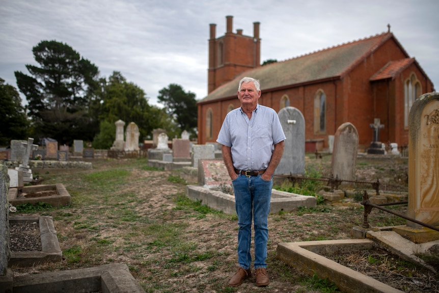 An older gentleman in jeans and blue button up shirt stands in a cemetery with a red brick church in the background.