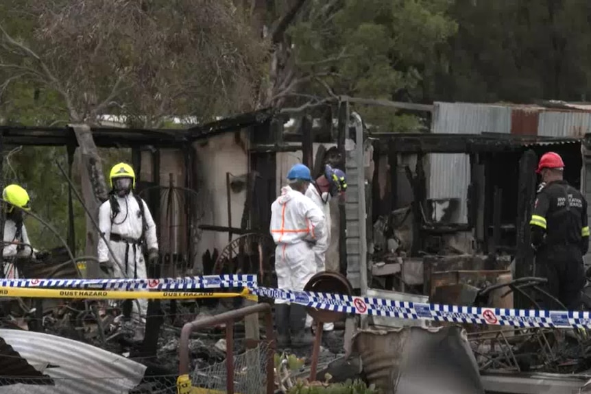 Men in hazmat suits and hard hats amid the charred ruins of a house on a country property.