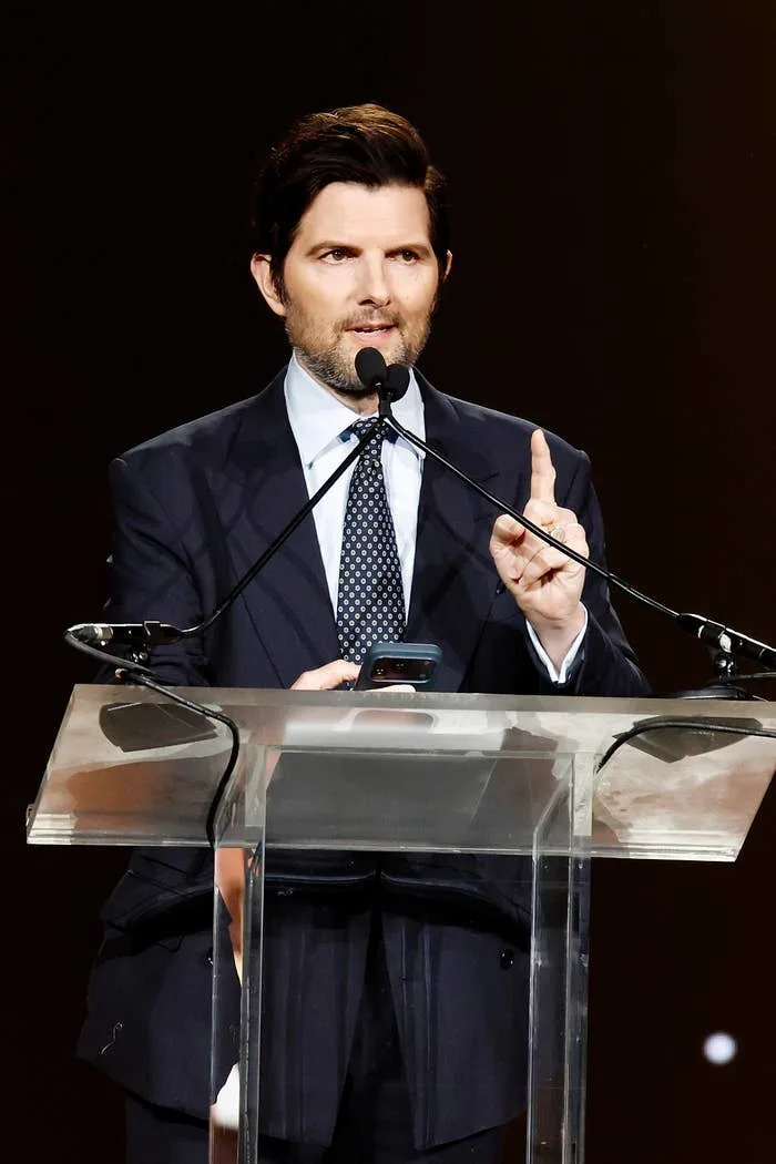 Adam Scott in a formal suit speaks at a podium, gesturing with one finger, during an event