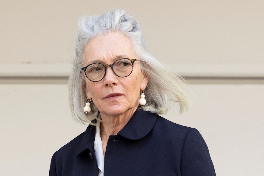 Prudence Murdoch walks outdoors in front of a white wall.